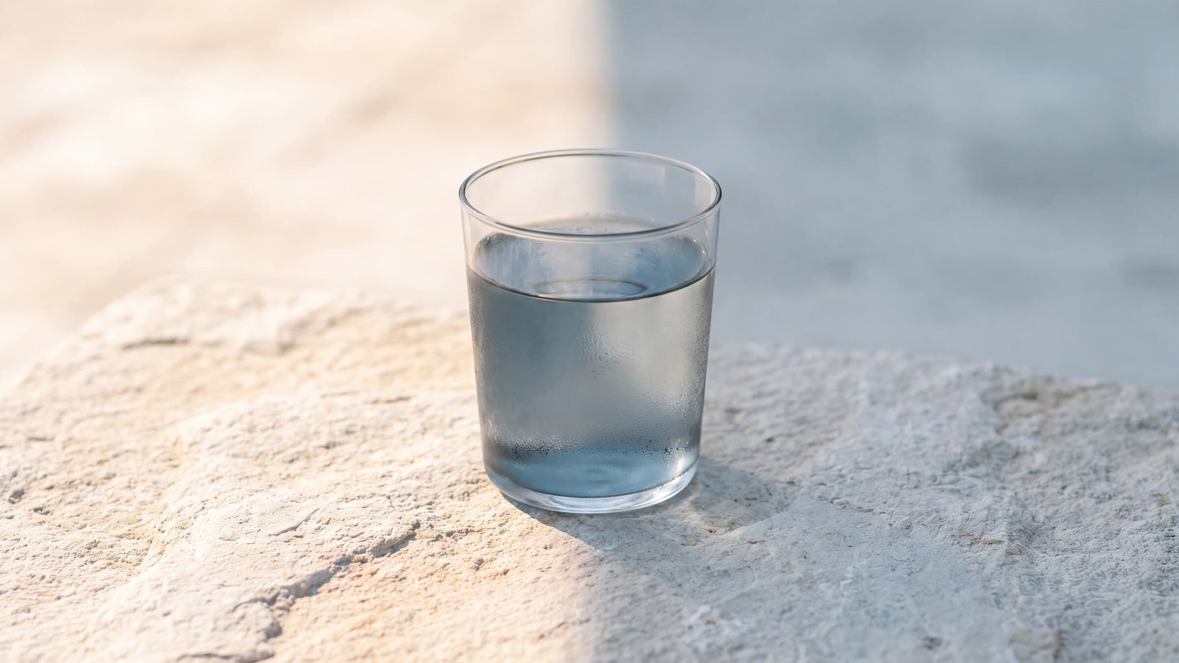 A glass of clear water on pale stone between warm sunlight and cool shade