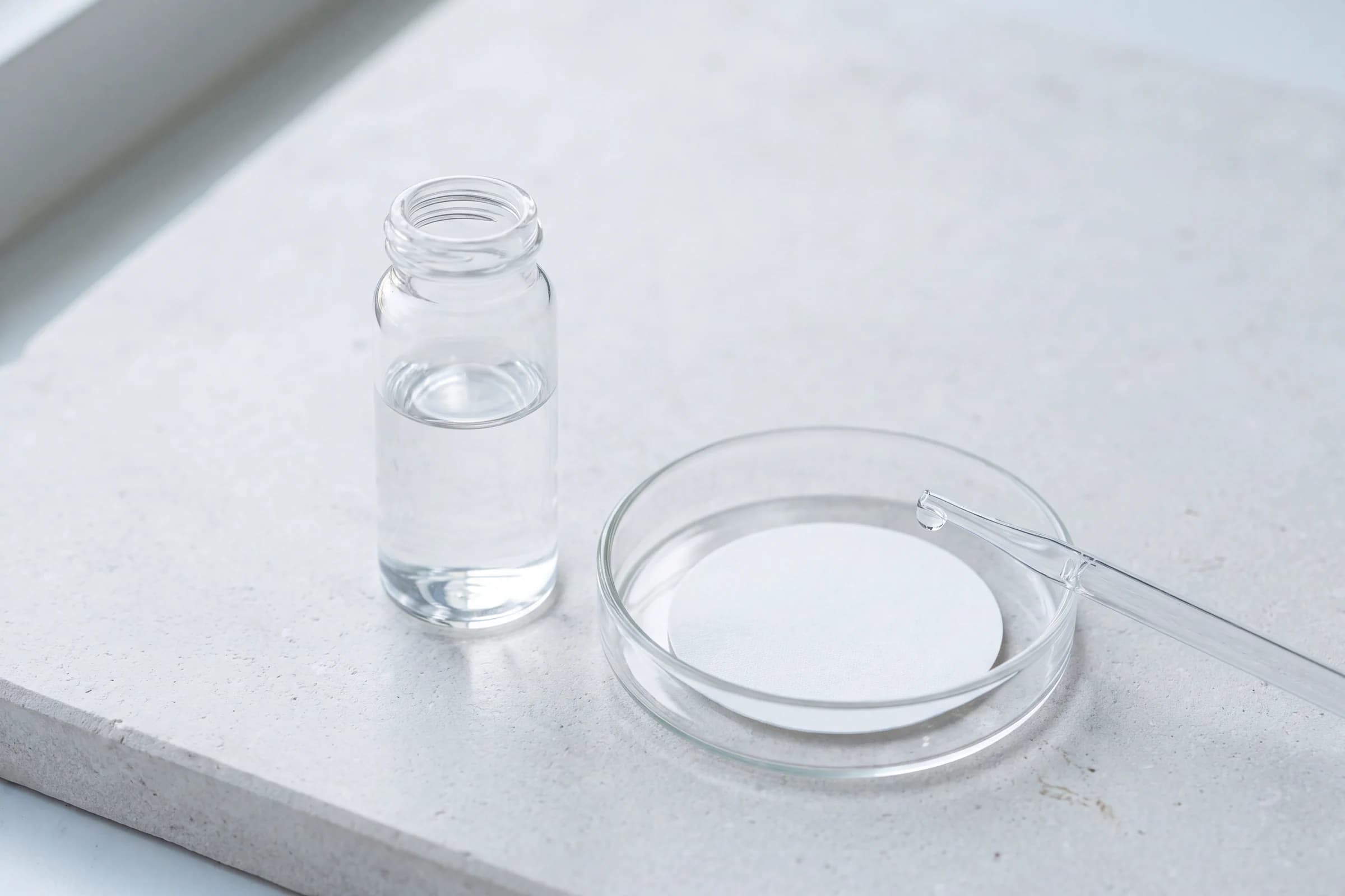 A clear water sample vial beside a white membrane filter in a glass dish and a pipette on pale stone